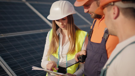 Ecological station. Alternative energy. Engineers use drawings on the solar farm in special uniform standing look around discuss the installation of sunny batteries. Solar panels fieldの写真素材