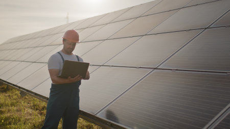 Portrait of happy male engineer in protective helmet with a laptop in his hands while looking to camera. Handsome man in uniform smiling while standing at solar power farm. Concept of green energyの写真素材