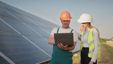 Technician and investor walking in Solar cell Farm through field of solar panels checking the panels at solar energy installation. Man holding laptop in her hands standing on field with solar panels.の写真素材