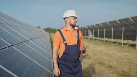 An employee of a power plant transmits commands by walkie-talkie. Happy caucasian adult engineer in helmet smiling at camera at solar power station outside. Ecological industry. Business teamの写真素材