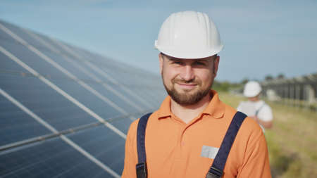 Happy caucasian adult engineer in helmet smiling at camera at solar power station outside. Ecological industry. Solar farm concept. An employee of a power plant transmits commands by walkie-talkieの写真素材