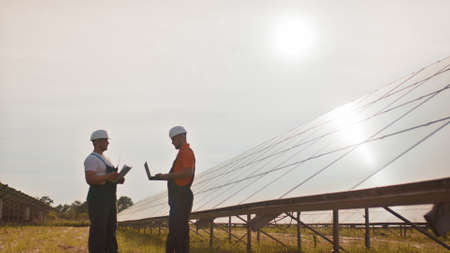 Coworking engineers with tablets on solar plant. Adult men in hardhats using tablets while standing outdoors on transformer platform. Transportation of clean energy. Solar park. Alternative energyの写真素材