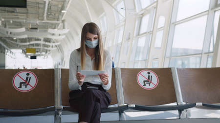 Confident business woman in a mask sitting at the airport, holding a passport and plane tickets, keeps a distance in a pandemic. Waiting at the airport. Travel concept. Woman looking at ticketsの写真素材