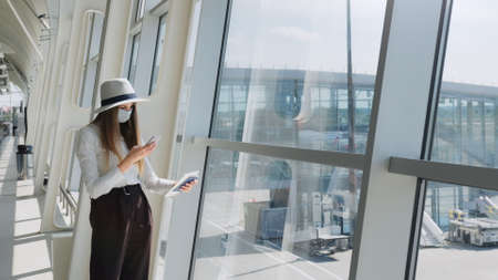 Young girl, business woman standing by the window in a modern airport holding tickets and a phone, looking at the camera. Portrait of a beautiful girl in a mask. Business trip by planeの写真素材