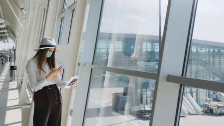 Young girl, business woman standing by the window in a modern airport holding tickets and a phone, looking at the camera. Portrait of a beautiful girl in a mask. Business trip by planeの写真素材