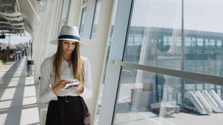 Elegant beautiful girl holding travel tickets and using a phone, happy girl looking at the camera in the airport terminal. Fashion woman portrait. Airport terminal. Business tripの写真素材