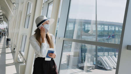 Portrait of a little girl talking on the phone near the airport terminal. Business trip concept. Young woman in a protective face mask standing in the airport hall by the window and uses smartphone.の写真素材