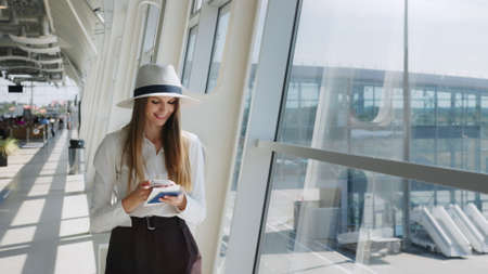 Elegant beautiful business girl holding travel tickets and using phone, happy girl looking at camera in airport terminal. Fashion woman portrait. Airport terminal. Business tripの写真素材