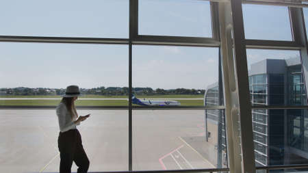 Young woman traveler, leisure or business standing at the glass window of a large international airport hub with a phone in his hands, scrolling the application for smartphones. Background of planeの写真素材