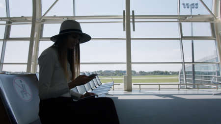 Elegant beautiful unrecognizable business girl sitting at the airport, holding a phone and tickets, slowly approaching the airport window. Airplane runway in the backgroundの写真素材