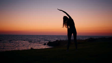 Young woman with slender body practising in fitness exercises during evening time near ocean. Silhouette of active healthy female. Regular training for body care. Woman fitness near ocean.の写真素材