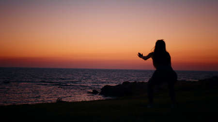 Fitness woman in silhouette doing squats exercises on beach near ocean. Young female in sportswear spending evening time for outdoors activity. Amazing colorful sunset on background. Fitness woman.の写真素材