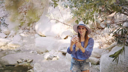 Attractive young woman in summer hat and clothes taking selfie on modern smartphone while standing outdoors. Female travel blogger making content to social network during vacation.の写真素材