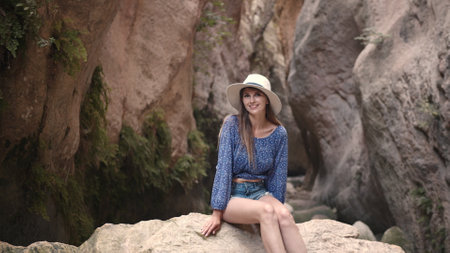 Stunning young woman sitting on a large rock among the rocks and smiling at the camera. Pretty Caucasian woman wearing summer hat and clothes. Enjoy summer vacationの写真素材