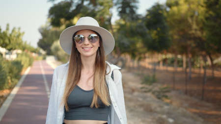 Portrait of a beautiful young girl in glasses. Happy young woman in summer clothes, hat and sunglasses walking with backpack outdoors. Palm trees around. Concept of people, vacation and tourism.の写真素材