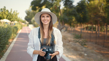 Happy young woman using modern digital camera for taking photos while walking. Female tourist in summer hat and clothes saving memories from trip. Young woman photographer wanderer.の写真素材