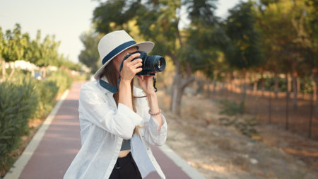 Happy young woman using modern digital camera for taking photos while walking. Female tourist in summer hat and clothes saving memories from trip. Young woman photographer wanderer.の写真素材
