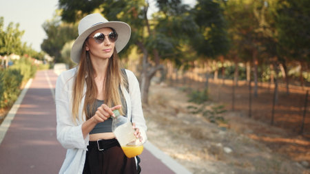 Pretty caucasian woman in white hat and sunglasses standing outdoors and drinking juice from bottle. Female tourist refreshing during warm sunny day on street.の写真素材