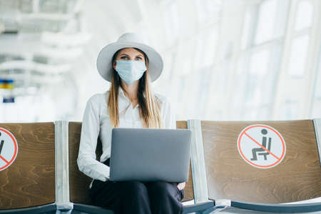 Woman in medical mask sitting at the airport terminal with a laptop. Young girl is waiting for departure at the gate and working online.の写真素材