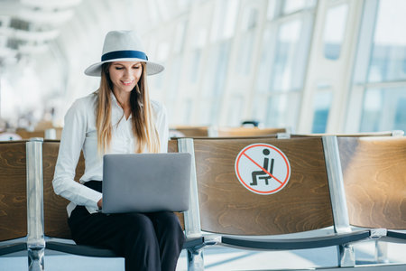 Departure lounge with a woman typing on a laptop. Girl tourist freelancer works and waits for flight in waiting room. Concept travel, remote work. Silhouette against the background of a large window.の写真素材