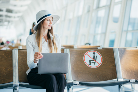 Business lady sitting at the airport on the background of a large window with a laptop on her lap, finishes work and approaches the window. Freelancer, The concept of work remotely from the officeの写真素材