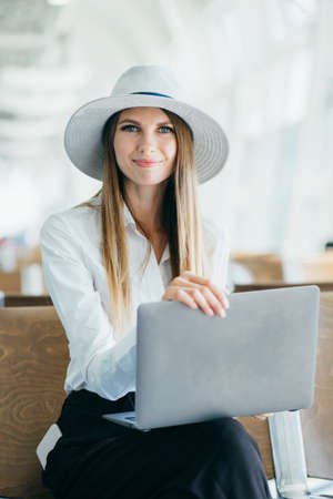 Young pretty woman printing on laptop sitting at airport. Girl tourist freelancer works and waits for flight in waiting room. Concept travel, remote work. Airport lounge with a lady using a laptop.の写真素材