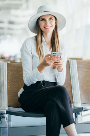 Girl uses smartphone in the airport lounge. Beautiful young girl using smartphone communication application airport arriving in new country. Woman travels around world. Smiling at the cameraの写真素材
