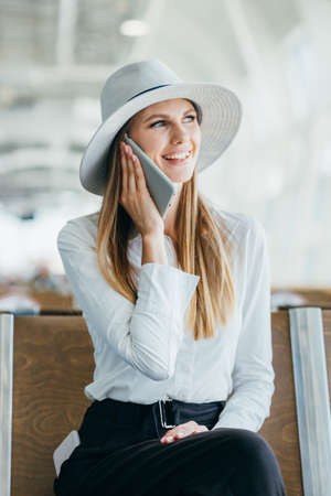 Portrait of a little girl talking on the phone near the airport terminal. Business trip concept. Young woman in a protective face mask standing in the airport hall by the window and uses smartphone.の写真素材