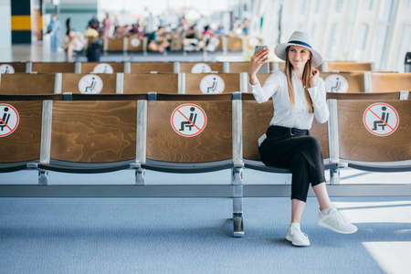 Young happy woman sitting at the gate and using smartphone while waiting for a flight at the airport. Travel concept. Travel passenger girl chatting and smiling at terminal departure loungeの写真素材