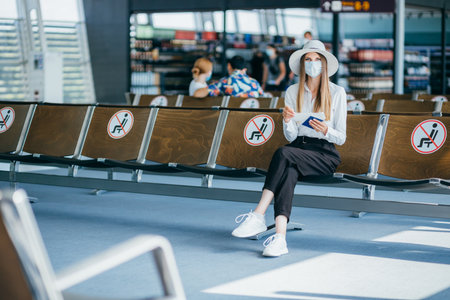 Young woman tourist wearing face mask holding passport and ticket, sitting in airport or train station on empty marked chairs under new normal and social covid distance regulations.の写真素材