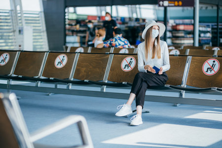 Woman tourist with mask with flight tickets and sitting in a socially distanced chair at the airport during the coronavirus epidemic. Concept of new normality of air transportation due to pandemic.の写真素材
