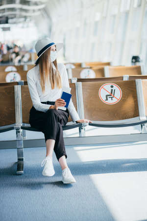 Confident business woman in a mask sitting at the airport, holding a passport and plane tickets, keeps a distance in a pandemic. Waiting at the airport. Travel concept. Woman looking at tickets.の写真素材
