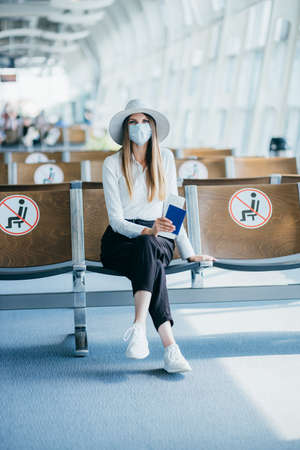Young woman in a mask sits in an airport lounge during a pandemic and shows her passport and ticket to the camera.の写真素材