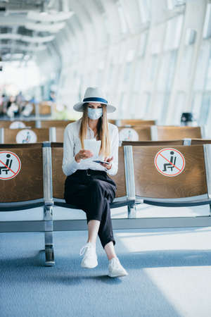 Confident business woman in a mask sitting at the airport, holding a passport and plane tickets, keeps a distance in a pandemic. Waiting at the airport. Travel concept. Woman looking at tickets.の写真素材