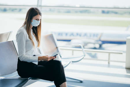 Happy smiling woman in protective mask holding boarding ticket and mobile while looking out the window in the airport.の写真素材