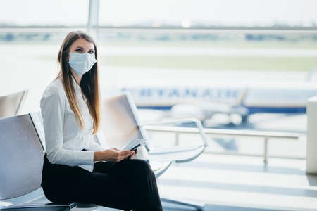 Young girl, business woman standing by the window in a modern airport holding tickets and a phone, looking at the camera. Portrait of a beautiful girl in a mask. Business trip by plane.の写真素材