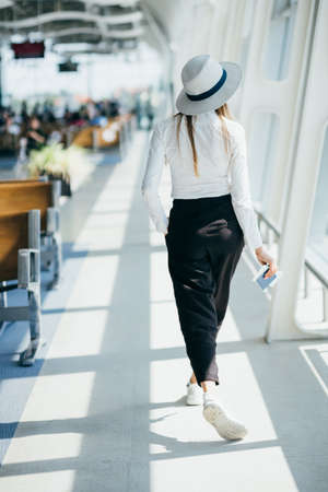 Young woman near window in an airport lounge waiting for flight aircraft. woman at the airport stands with tickets, overall plan.の写真素材