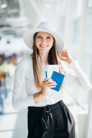 Cheerful businesswoman waiting for her flight at airport. Young woman at the airport. Young woman holding passport and boarding pass at airport. Attractive young woman going on a trip by air.の写真素材