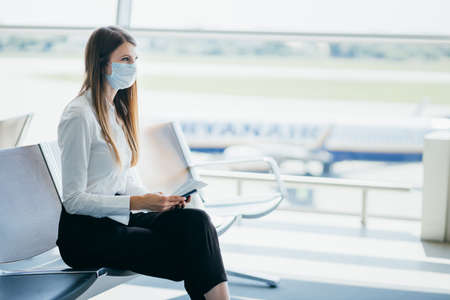 Young woman traveler stylishly dressed, leisure or business sitting by the glass window of a large international airport hub with a phone in hand, scrolling the smartphone appの写真素材
