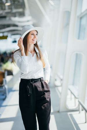 Cool young woman in boater and white dress smiles and looks into camera outside. Tourist holds passport and poses on crosswalk near airport.の写真素材
