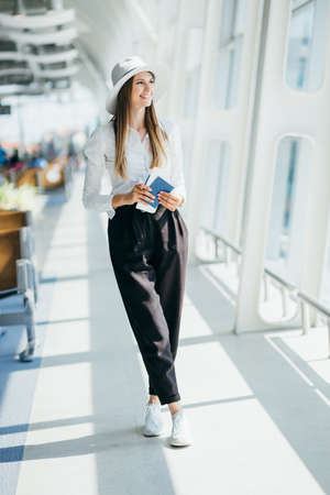 Elegant beautiful business girl holding travel tickets and using phone, happy girl looking at camera in airport terminal. Fashion woman portrait. Airport terminal. Business trip.の写真素材