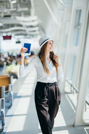 Elegant beautiful girl holding travel tickets and using a phone, happy girl looking at the camera in the airport terminal. Fashion woman portrait. Airport terminal. Business trip.の写真素材