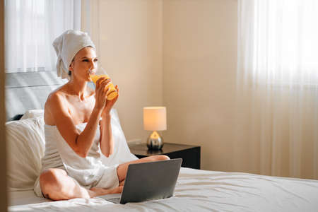 Young girl with long hair in white robe using laptop having breakfast orange juice in bed of hotel room. Lovely woman sitting, stretch lazily and using laptop computer and drinking orange juice.の写真素材