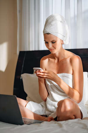 Pretty woman wrapped in bath towel lying on bed with laptop on knees and holding smartphone in hands. Lifestyles with modern technology.の写真素材
