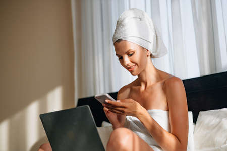 Pretty woman wrapped in bath towel lying on bed with laptop on knees and holding smartphone in hands. Lifestyles with modern technology. A woman works on vacation at the hotel, after a bath or shower.の写真素材