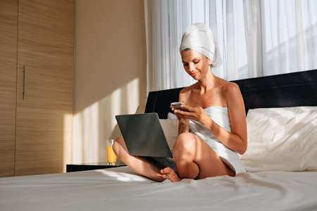 Pretty woman wrapped in bath towel lying on bed with laptop on knees and holding smartphone in hands. Lifestyles with modern technology. A woman works on vacation at the hotel, after a bath or shower.の写真素材