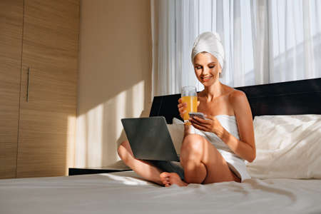 Woman wrapped in a bath towel lying in bed with a laptop on her lap and holding a smartphone in her hands. Lifestyle with modern technologies. Young girl with a laptop talking on the phone and smilingの写真素材