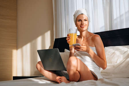 Caucasian young woman typing on laptop while sitting on bed with fresh fruits and juice. Happy lady enjoying vacation time at cozy hotel room. Woman typing on laptop while sitting on bed with fruits.の写真素材