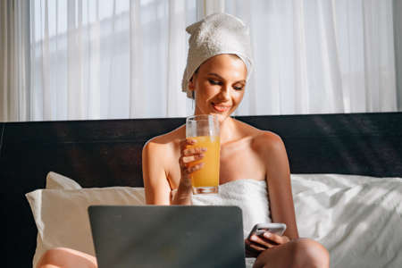 Woman have breakfast in modern hotel apartment. Window light portrait young girl in bathrobe lying on bed using laptop for work. Out of focus tray with meal, juice, plate, teapot on background.の写真素材