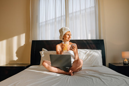Side view of happy woman typing on wireless laptop and drinking orange juice from glass at hotel room. Caucasian female sitting on comfy bed with crossed legs, with bath towel on head and bodyの写真素材